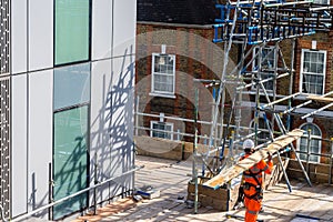 A scaffolder carries a wooden plank on a building site, wearing orange hi-vis protection clothes. photo