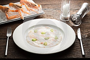 Scallops carpaccio served with chili, langoustines on a white plate and wooden rustic brown table with dinner settings. Close up