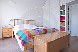 Scandi style bedroom interior with wooden bedroom furniture, white painted walls, white bedding and colourful blanket.