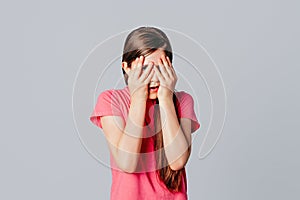 Scared anxious young girl amazed shocked hands cover face, wear in casual pink t shirt, standing over gray background. Young