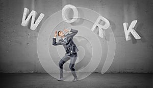 A scared businessman hides under large concrete letters making a word Work above him on a grey background.