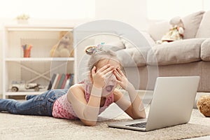 Scared casual little girl watching movie on laptop while lying on the floor at home