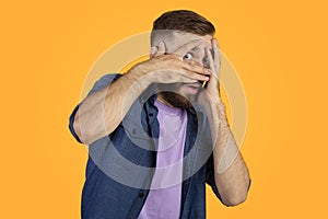 Scared Caucasian guy hiding behind his hands, peeping between fingers on orange studio background