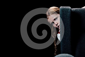 Scared child looking at camera while hiding behind armchair