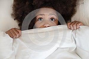 Scared african kid looking at camera covering blanket in bed photo