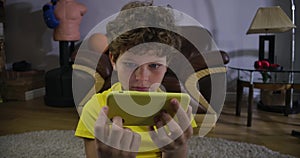 Scared face of Caucasian boy watching movies on smartphone screen. Cute child with grey eyes and curly hair absorbed by