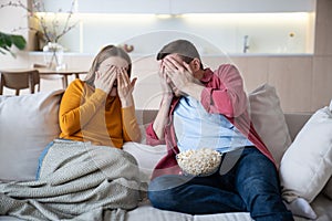 Scared family couple cover eyes with palms watching horrifying movie series sitting on couch at home