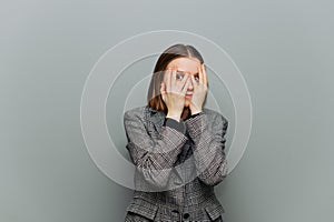 Scared female worker in jacket isolated on gray background, looking through fingers on face with shocked face