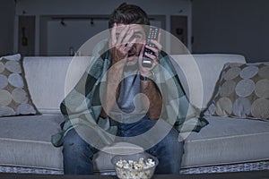 Scared and funny man alone at night in living room couch watching horror scary movie or scary show in television eating popcorn
