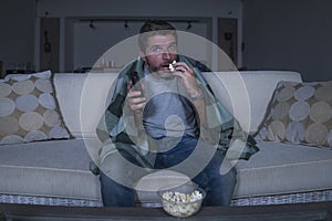 Scared and funny man alone at night in living room couch watching horror scary movie or scary show in television eating popcorn