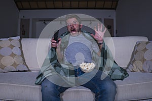 Scared and funny man alone at night in living room couch watching horror scary movie in television screaming and eating popcorn