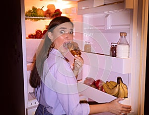 Scared girl in pajamas eating croissant near opened fridge