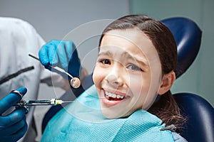 Scared girl sit in dental chair in room. She afraid of dentist and teeth pain. Adult hands hold tools.