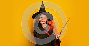 Scared girl in wizard student costume with magic wand holding hand on face and looking at camera on yellow background.