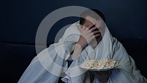 Scared handsome caucasian man watching horror movie late at night, eating popcorn with blanket, man watching horror