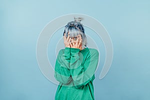 Scared hipster girl in a green sweatshirt and with blue hair stands on a blue background and covers her face in fear, looking at