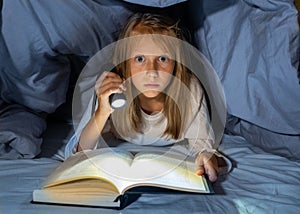 Scared little girl reading scary book under bed cover holding a flashlight late at night