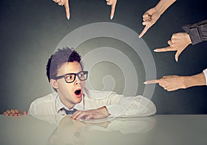 Scared man employee hiding under the table being accused by people who point fingers at him