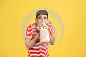 Scared nervous man in striped t-shirt hardly breathing using paper bag and looking at camera with big shocked eyes, trying to calm