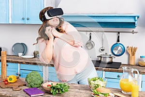 scared overweight woman in virtual reality headset standing at table with fresh vegetables in kitchen