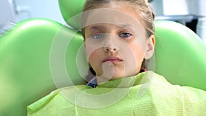 Scared preschool girl frowns and sits in dentist chair waiting for procedure