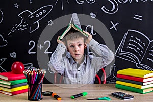Scared and puzzled schoolboy sitting at the desk holding the book above the head, surrounded with school supplies