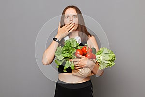 Scared shocked Caucasian woman holding organic vegetables standing isolated over gray background covering mouth with palm looking