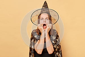 Scared shocked frighten woman wearing witch costume and cone hat isolated over beige background screaming with eyes full of fear