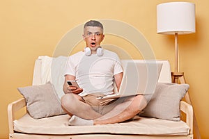 Scared shocked surprised man wearing white T-shirt and shorts sitting on sofa against beige background looking at camera with big