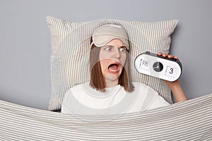 Scared shocked woman in white T-shirt and sleeping eye mask lie in bed on pillow under blanket isolated on gray background being