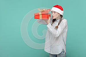 Scared teenager girl in Santa Claus hat, holding present box, looking inside with big eyes.