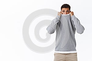 Scared, timid and cute young guy feeling afraid of horror movies, pulling sweater on face and frowning with arched sad