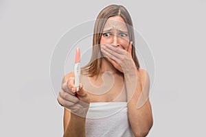 Scared and worried young woman stands and looks at pregnancy test. She holds it in hands. Model coveres mouth with hand