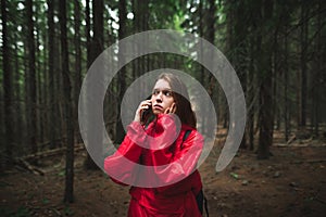 Scared young female tourist in red raincoat is lost in forest calling someone on the phone, touching her face. Terrified traveler