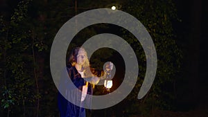 Scared young girl holding oil lamp and looking at something in dark forest at night