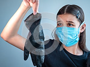 Scared young girl wearing a blue medical facemask holds a black latex glove by finger tips in streched out arm. Covid-19 concept