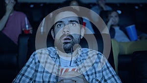 Scared young man watching horror film in cinema holding glass with drink