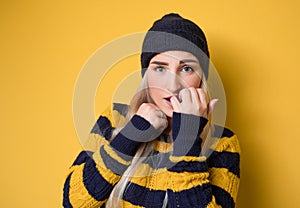 Scared young woman biting fingernail,being terrible accident, model wearing woolen cap and sweater, isolated on yellow background