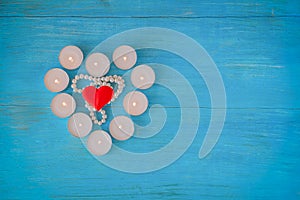 A scarlet heart on pearl beads is surrounded by burning small heart-shaped candles on a blue wooden background