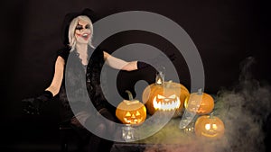 Scary beautiful girl witch laughs, taunts, gloats, celebrates halloween with funny glowing burning pumpkins in smoke