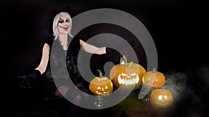 Scary beautiful girl witch laughs, taunts, gloats, celebrates halloween with funny glowing burning pumpkins in smoke