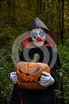 Scary clown in the forest with a pumpkin for Halloween.