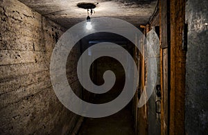 A scary dark concrete corridor in the basement, lit by a single light bulb hanging from the low ceiling. Copy space