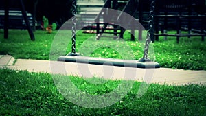 Scary empty swing with chains swaying at playground for child, moved from wind, on green meadow background