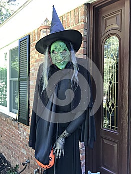 The scary green face witch with black outfit and pointed hat in front of the house