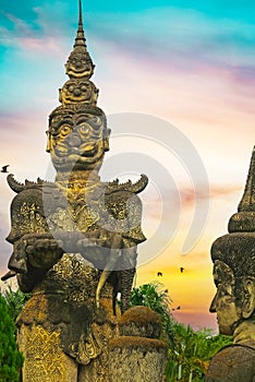 Scary mythological hindu mystic creature stone sculpture and buddha head, dramatic sunset sky, palm trees- Buddha Park Wat Xieng