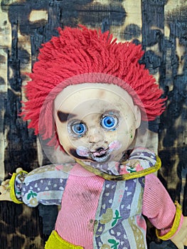 scary and old battered clown doll with burnt face and red hair on the background of burnt wooden board