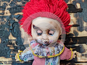 scary and old battered clown doll with burnt face and red hair on the background of burnt wooden board