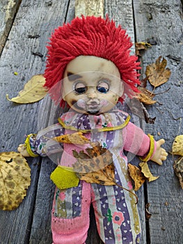 scary and old battered clown doll with burnt face and red hair on the background of old wooden board with autumn leaves