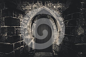 Scary pointy wooden door in an old and wet stone wall building with cross, skull and bones at both sides in black and white.